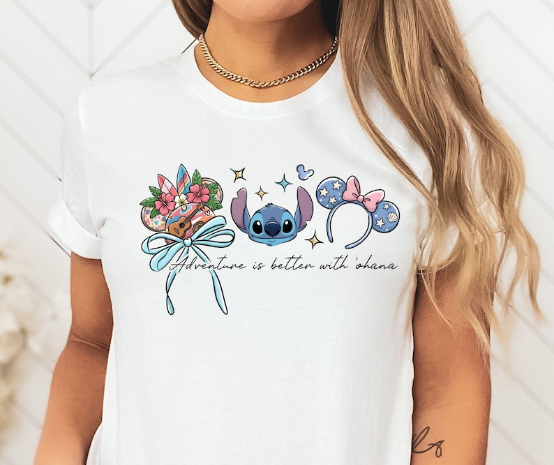 May include: White t-shirt featuring a colorful graphic of Stitch, Mickey Mouse ears, and a ukulele. The design includes the phrase "Adventure is better with 'ohana" in a script font. The design is in shades of blue, pink, and brown.