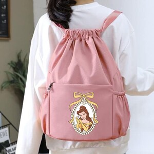 May include: A pink drawstring backpack with a front pocket and side pockets. The front of the backpack features an oval portrait of a cartoon character in a yellow dress. The backpack has pink straps.