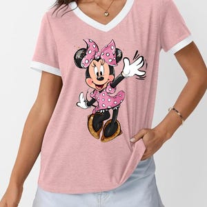 K&ouml;nnte beinhalten: Rosafarbenes V-Ausschnitt-T-Shirt mit wei&szlig;em Besatz und Minnie Mouse-Grafik. Minnie tr&auml;gt ein rosa gepunktetes Kleid und eine Schleife mit goldenen Schuhen. Das Shirt hat einen l&auml;ssigen Stil.