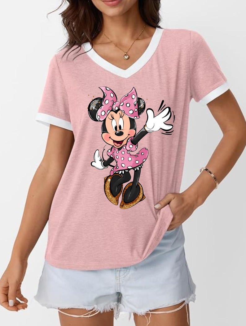 K&ouml;nnte beinhalten: Rosafarbenes V-Ausschnitt-T-Shirt mit wei&szlig;em Besatz und Minnie Mouse-Grafik. Minnie tr&auml;gt ein rosa gepunktetes Kleid und eine Schleife mit goldenen Schuhen. Das Shirt hat einen l&auml;ssigen Stil.