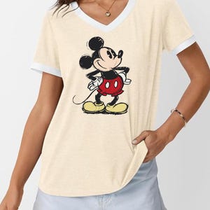 May include: Cream-colored V-neck t-shirt with white trim featuring a classic Mickey Mouse graphic. The cartoon character is depicted in his signature red shorts, yellow shoes, and black gloves, with his hands on his hips.