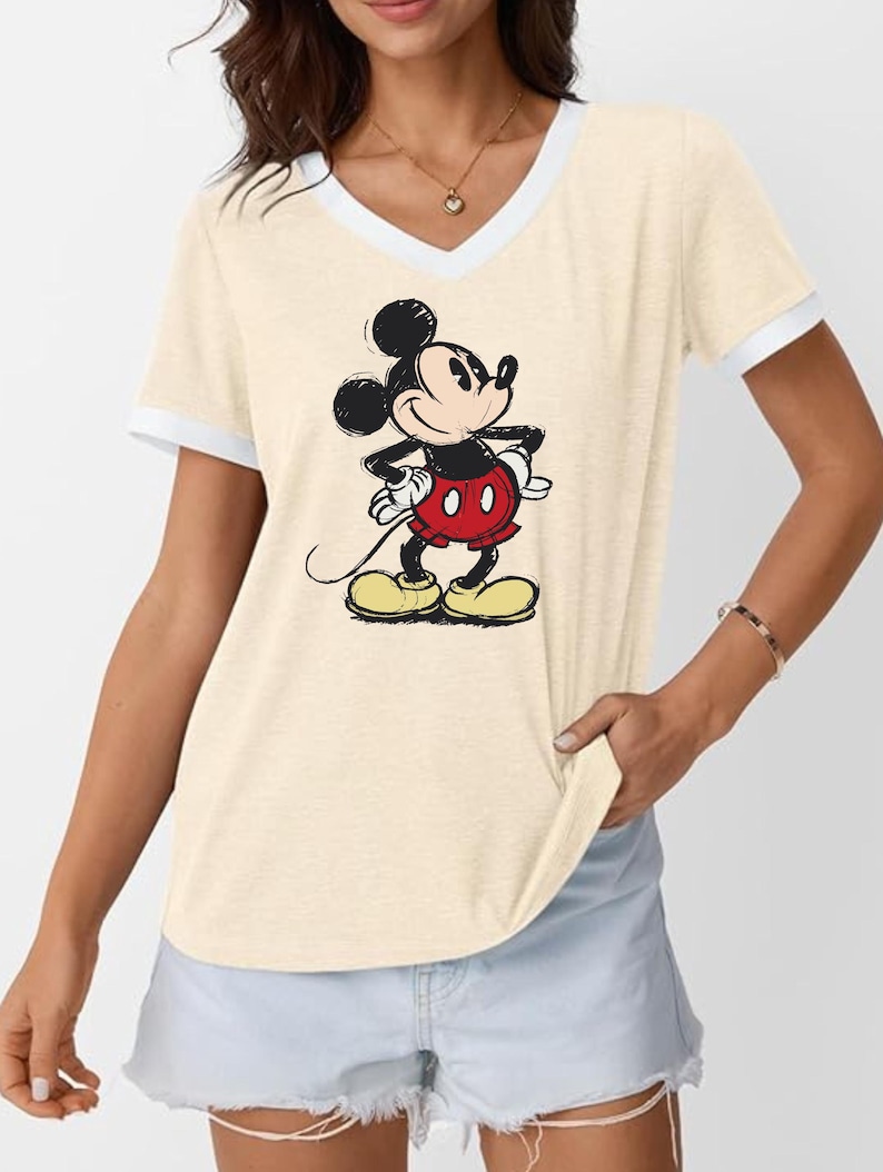 May include: Cream-colored V-neck t-shirt with white trim featuring a classic Mickey Mouse graphic. The cartoon character is depicted in his signature red shorts, yellow shoes, and black gloves, with his hands on his hips.