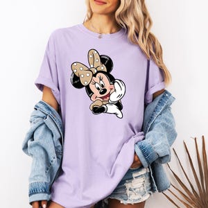 May include: A lilac t-shirt with a cartoon Minnie Mouse design. The graphic shows Minnie Mouse with a polka-dot bow, black ears, and a cheerful expression. The top has short sleeves and a loose fit, ideal for everyday wear.