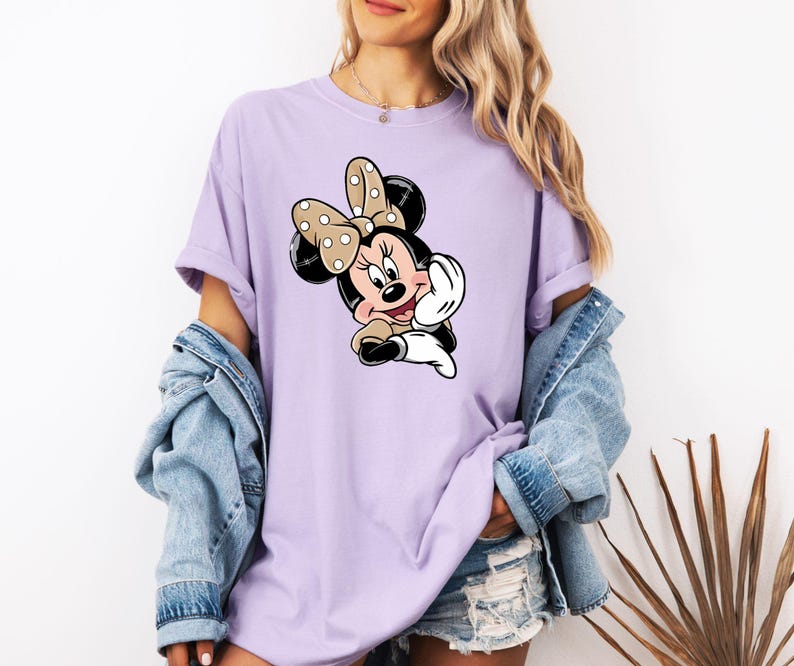 May include: A lilac t-shirt with a cartoon Minnie Mouse design. The graphic shows Minnie Mouse with a polka-dot bow, black ears, and a cheerful expression. The top has short sleeves and a loose fit, ideal for everyday wear.
