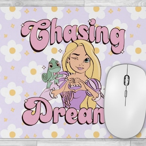 Peut inclure: Tapis de souris rectangulaire avec une illustration de dessin animé d'une femme blonde faisant un clin d'œil et formant un cœur avec ses mains. Un caméléon vert est sur son épaule. Les mots "Chasing Dreams" sont écrits en rose.