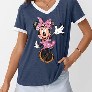 K&ouml;nnte beinhalten: Marineblaues V-Ausschnitt-T-Shirt mit wei&szlig;em Besatz und einem Cartoon-Bild von Minnie Mouse. Minnie Mouse tr&auml;gt ein rosa gepunktetes Kleid, eine Schleife und Schuhe. Das Shirt ist ein l&auml;ssiger Stil.