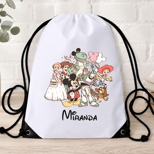 May include: A white drawstring backpack featuring a colourful graphic print of Disney characters including Mickey Mouse, Woody, Buzz Lightyear, Jessie, Rex, and Bo Peep. The name Miranda is printed in black script below the character illustration.