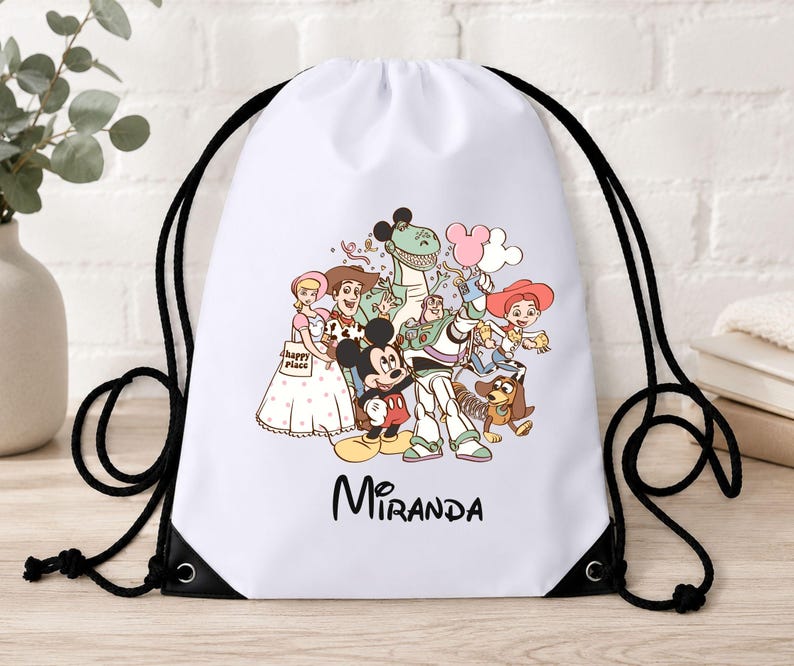 May include: A white drawstring backpack featuring a colourful graphic print of Disney characters including Mickey Mouse, Woody, Buzz Lightyear, Jessie, Rex, and Bo Peep. The name Miranda is printed in black script below the character illustration.