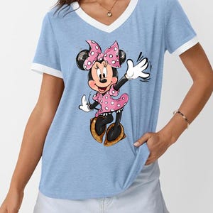 K&ouml;nnte beinhalten: Hellblaues V-Ausschnitt-T-Shirt mit wei&szlig;em Besatz und Minnie Mouse-Grafik. Minnie tr&auml;gt ein rosa gepunktetes Kleid und eine Schleife mit goldenen Glitzerschuhen. Ein l&auml;ssiger Stil.