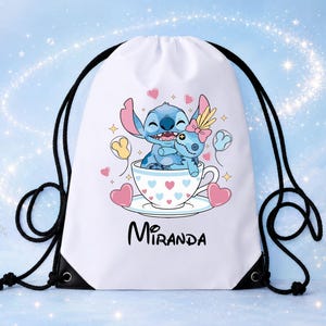 Op de afbeelding: Een witte rugzak met trekkoord voorzien van een afbeelding van een blauw alien-personage dat een pop knuffelt in een theekopje versierd met roze en blauwe hartjes. De naam Miranda staat in zwarte letters onder het kopje, met zwarte koorden.