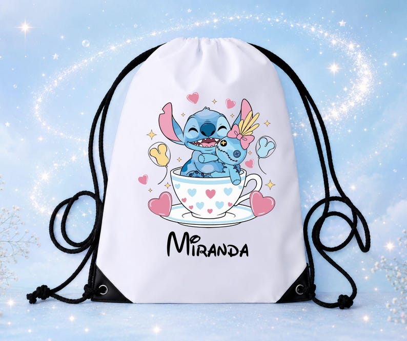 Op de afbeelding: Een witte rugzak met trekkoord voorzien van een afbeelding van een blauw alien-personage dat een pop knuffelt in een theekopje versierd met roze en blauwe hartjes. De naam Miranda staat in zwarte letters onder het kopje, met zwarte koorden.