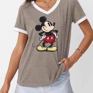 May include: A gray V-neck t-shirt with white trim on the collar and sleeves. The shirt features a graphic of Mickey Mouse in his classic outfit: red shorts, yellow shoes, and white gloves. The cartoon character is standing with his hands on his hips.