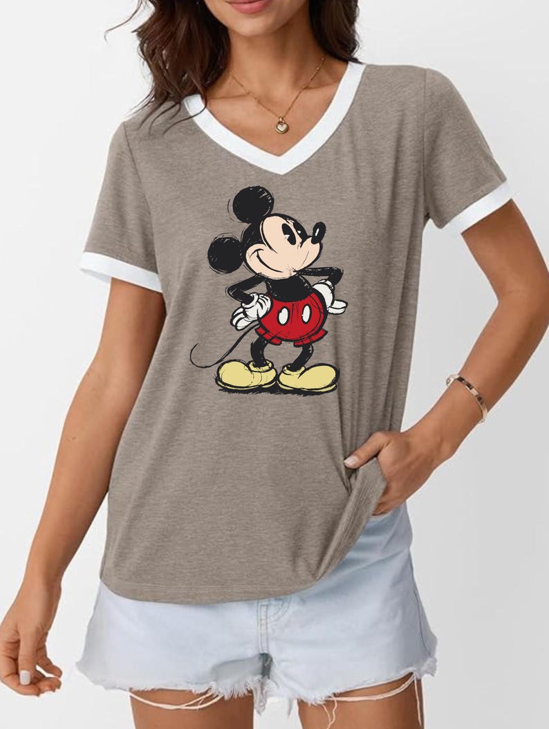 May include: A gray V-neck t-shirt with white trim on the collar and sleeves. The shirt features a graphic of Mickey Mouse in his classic outfit: red shorts, yellow shoes, and white gloves. The cartoon character is standing with his hands on his hips.