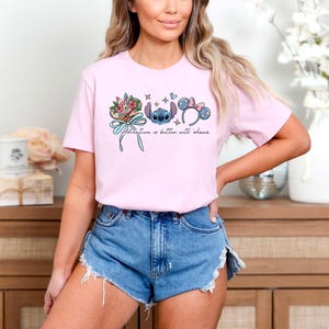 May include: A light pink t-shirt featuring a graphic of Stitch, floral elements, and Minnie Mouse ears. The text "Adventure is better with ohana" is printed below the graphic. The shirt is paired with denim shorts.