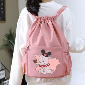 May include: Pink drawstring backpack featuring a Dumbo graphic. The backpack has a front zippered pocket and side pockets. The Dumbo illustration includes a black hat, red collar, and a brown Mickey Mouse glove. The straps are pink.
