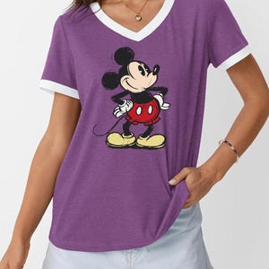 May include: Purple V-neck t-shirt with white trim featuring a graphic of Mickey Mouse. The cartoon character is in a classic pose, wearing red shorts, yellow shoes, and white gloves. The shirt is a casual style.
