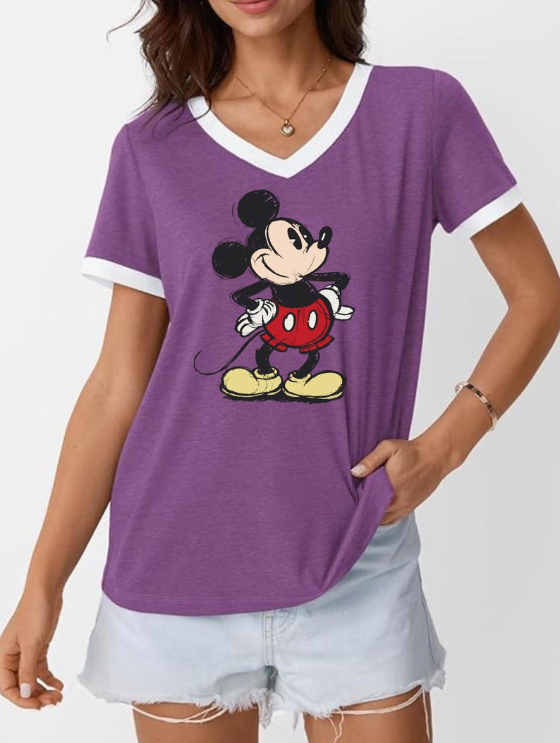 May include: Purple V-neck t-shirt with white trim featuring a graphic of Mickey Mouse. The cartoon character is in a classic pose, wearing red shorts, yellow shoes, and white gloves. The shirt is a casual style.