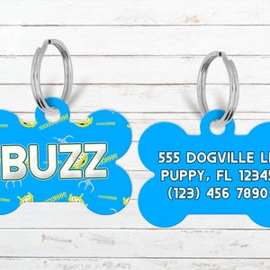 May include: Two blue dog bone-shaped pet tags. One tag has the name "BUZZ" in white block letters, with cartoon aliens and text. The other tag has an address and phone number in white text.