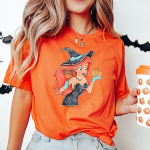 May include: Orange t-shirt featuring a cartoon illustration of a witch-themed character holding a pumpkin and a Mickey Mouse-shaped lollipop. The character has red hair and is wearing a black dress and witch hat. A white coffee cup with pumpkin graphics is also visible.