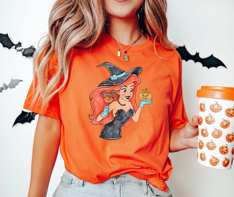 May include: Orange t-shirt featuring a cartoon illustration of a witch-themed character holding a pumpkin and a Mickey Mouse-shaped lollipop. The character has red hair and is wearing a black dress and witch hat. A white coffee cup with pumpkin graphics is also visible.
