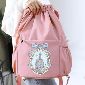 May include: Pink drawstring backpack with a front pocket and side pockets. The front features an oval design with a castle illustration, a blue bow, and a pearl border. The backpack has pink straps and a drawstring closure.