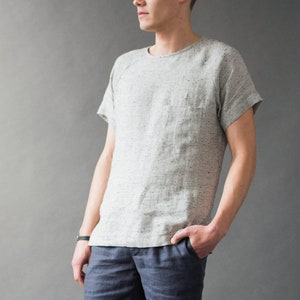 Pode incluir: Um homem vestindo uma camiseta de linho cinza claro com gola redonda e mangas curtas, shorts de linho azul e tênis altos azuis. Ele está em pé em frente a uma parede cinza com uma pequena planta de cacto em um vaso preto em um banquinho de metal preto.