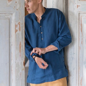 Op de afbeelding: Een man draagt een blauw linnen shirt met een knoopsluiting en lange mouwen. Het shirt heeft een trekkoord detail aan de zijkanten. Hij draagt een beige broek.
