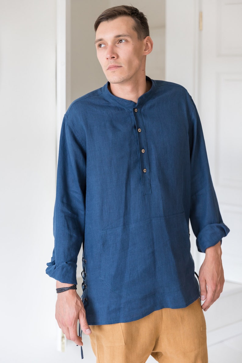 Op de afbeelding: Een blauw linnen overhemd met een mandarijnkraag en lange mouwen. Het overhemd heeft een knoopsluiting en een kantdetail aan de zijkant. Het model draagt het overhemd met een bruine broek.