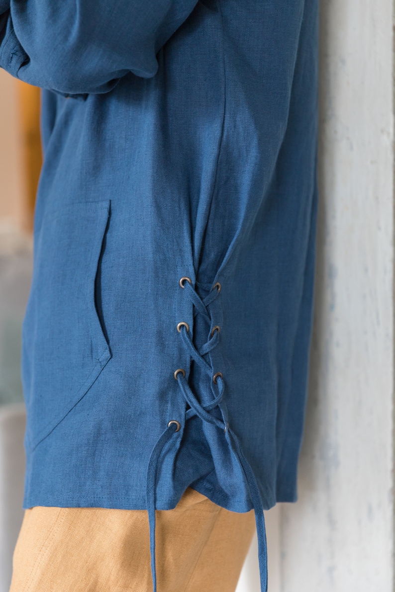 Op de afbeelding: Een blauw linnen shirt met een zijdelingse veterdetail. Het shirt heeft een zak aan de voorkant en is in de taille geknoopt.