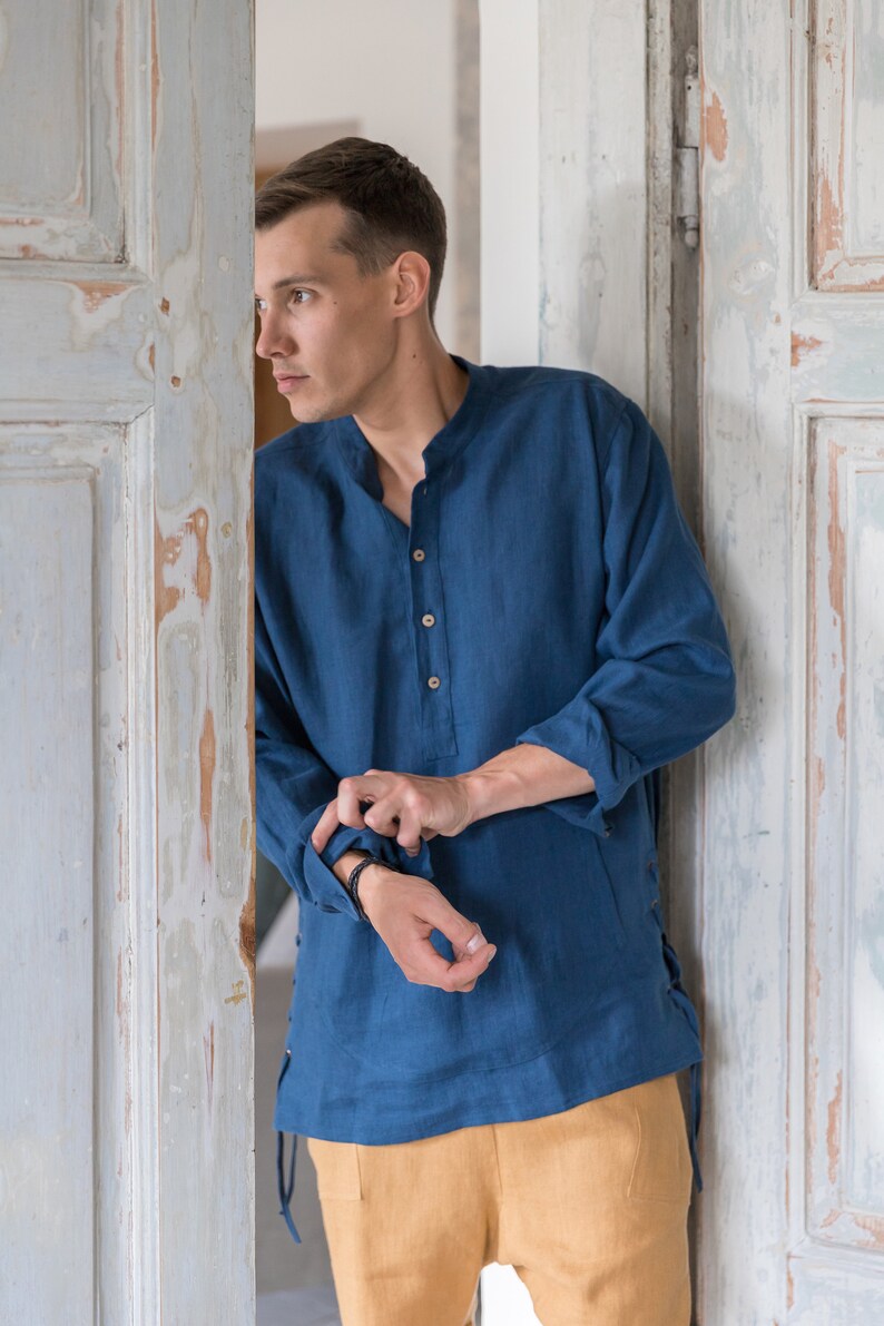 Op de afbeelding: Een man draagt een blauw linnen shirt met een knoopsluiting en lange mouwen. Het shirt heeft een trekkoord detail aan de zijkanten. Hij draagt een beige broek.