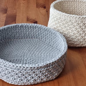 Peut inclure: Deux paniers faits à la main au crochet. L'un est un panier rond gris clair avec un profil bas. L'autre est un panier plus haut, blanc cassé, de forme cylindrique. Les deux paniers ont un aspect texturé et tissé et reposent sur une surface en bois.