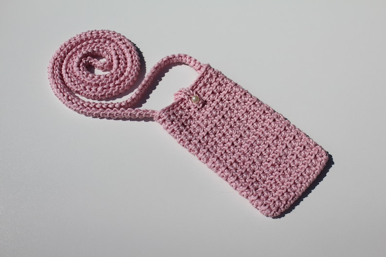 May include: A pink crocheted phone case with a long strap. The case has a pearl button closure.