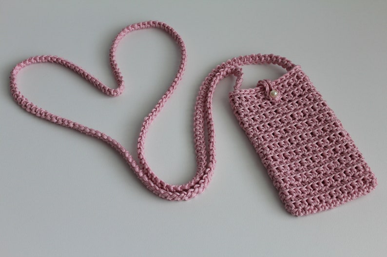 May include: A pink crocheted phone case with a pearl button and a long pink crocheted strap. The strap is shaped into a heart.