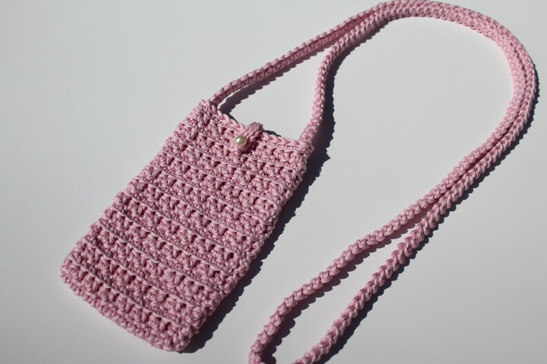 May include: A pink crocheted crossbody bag with a pearl accent. The bag is small and has a long strap.