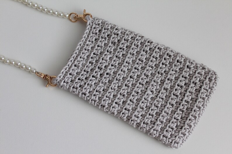 May include: A gray crocheted phone pouch with a pearl chain strap. The strap has gold clasps.