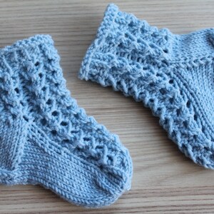 Può includere: Due calzini da neonato lavorati a maglia azzurri. I calzini presentano un motivo traforato strutturato sulla parte superiore e polsini e piedi a coste. I calzini sono realizzati con un filato morbido, forse lana.