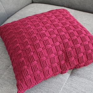 May include: A burgundy knitted cushion with a square pattern. The cushion is sitting on a grey sofa.