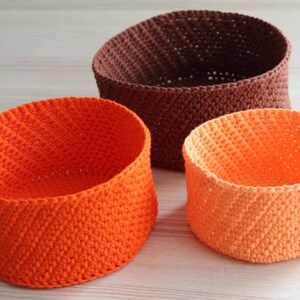Puede incluir: Tres cestas de crochet de diferentes tamaños. La cesta más grande es marrón, la cesta mediana es naranja y la cesta más pequeña es de un tono naranja más claro.