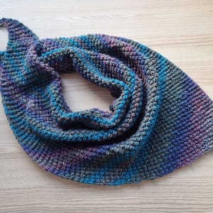 May include: A crocheted scarf in shades of blue, purple, and gray. The scarf is folded in a triangular shape and has a textured, woven pattern.