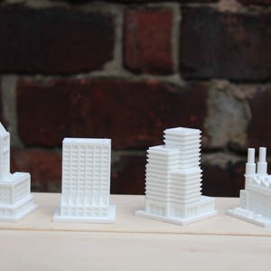 May include: Four miniature white architectural models of buildings, including a tower, a skyscraper, a multi-story structure, and a factory with smokestacks. The models are displayed on a light-colored wooden surface against a brick background.