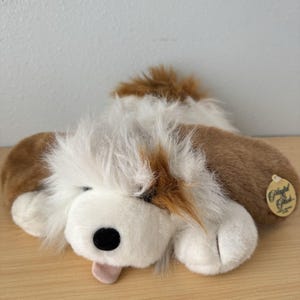 May include: A plush dog toy in shades of brown, white, and tan, with a black nose and pink tongue. The toy has a fluffy, shaggy appearance and a tag that reads "Playful Plush". The dog is lying down on a wooden surface.