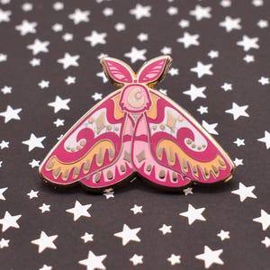 May include: A pink and gold moth-shaped enamel pin with intricate details. The pin features a detailed design with pink, gold, and cream colors. The background is black with white stars.