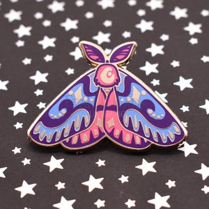 May include: A colorful moth enamel pin with a gold outline. The pin features a purple body and wings with blue, pink, and gold accents. The pin is set against a black background with white stars.