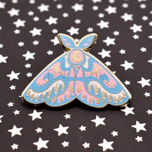 May include: A blue, pink, and gold moth-shaped enamel pin. The pin features a detailed design with a gold outline and a butterfly-like shape. The background is black with white stars.