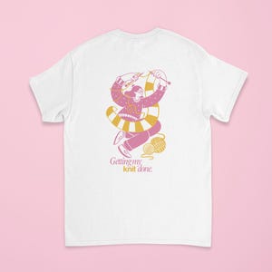 May include: White t-shirt featuring a pink and yellow illustration of a person knitting. The design includes a ball of yarn and the text "Getting my knit done." The graphic is in a playful, cartoon style.