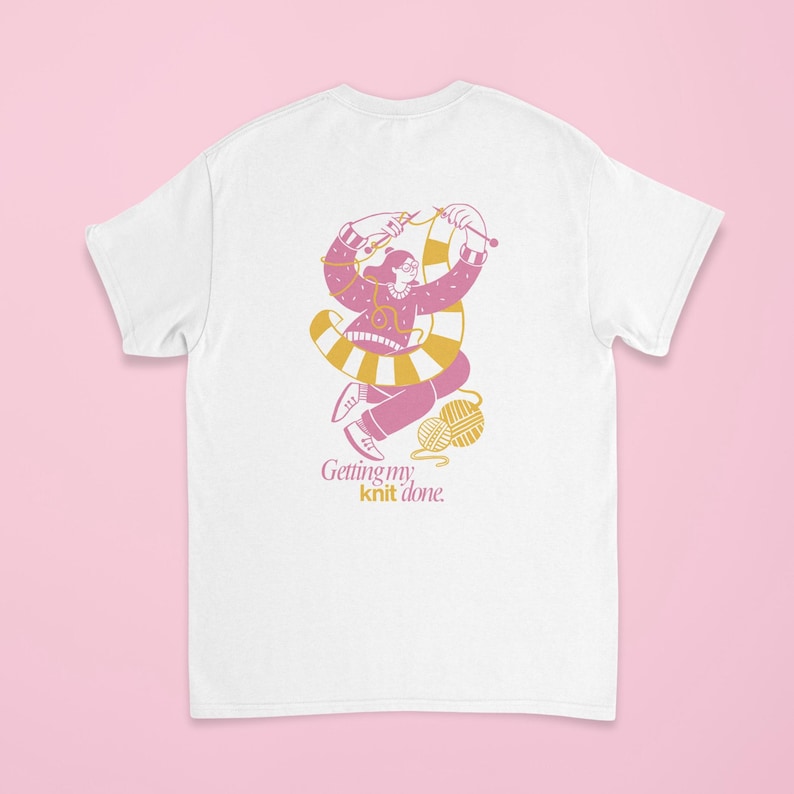 May include: White t-shirt featuring a pink and yellow illustration of a person knitting. The design includes a ball of yarn and the text "Getting my knit done." The graphic is in a playful, cartoon style.