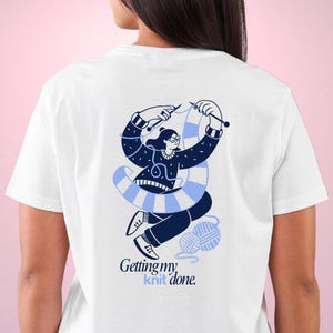 May include: White t-shirt with a blue illustration of a person knitting. The illustration shows a woman with glasses, knitting needles, and a blue and white striped scarf. The text "Getting my knit done" is below the illustration.