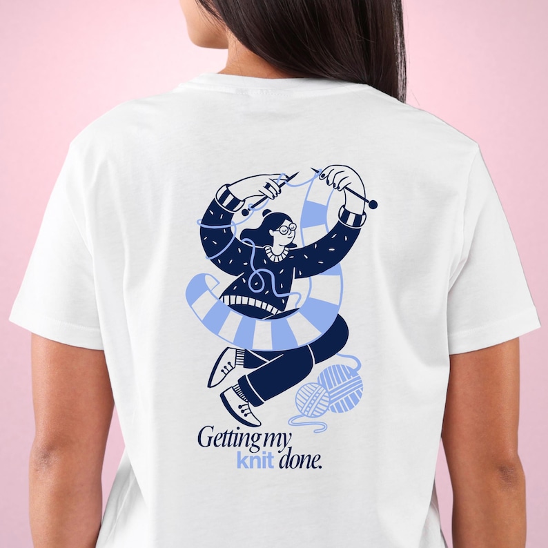 May include: White t-shirt with a blue illustration of a person knitting. The illustration shows a woman with glasses, knitting needles, and a blue and white striped scarf. The text "Getting my knit done" is below the illustration.