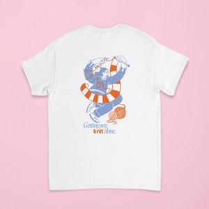 May include: White t-shirt featuring a blue and orange illustration of a person knitting. The illustration includes a ball of yarn and the text "Getting my knit done."