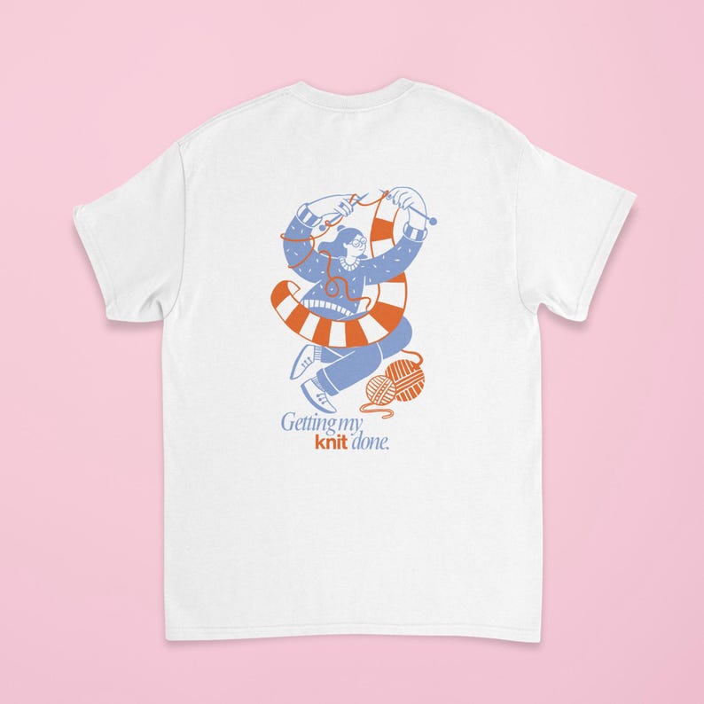 May include: White t-shirt featuring a blue and orange illustration of a person knitting. The illustration includes a ball of yarn and the text "Getting my knit done."